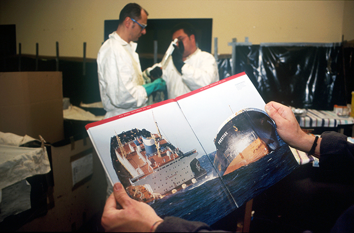 Un voluntario abre una revista con la foto del "Prestige" hundiéndose mientras otros dos se disponen a atender a la fauna petroleada.